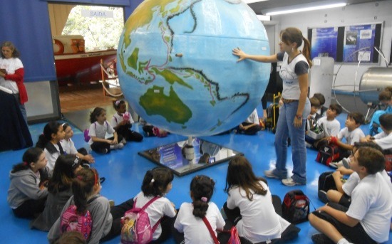 A foto ilustra um grupo de crianças sentadas em roda no chão do Museu de Oceanografia da USP, em volta de um globo terrestre grande, fixado pelo teto, acompanhando a explicação da monitora.