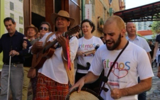 Na foto, o grupo composto pela equipe da Ritmos e do grupo Folias e Folguedos realiza um cortejo no corredor de entrada do Sesc Pompéia, cantando, tocando instrumentos e acompanhados pelos frequentadores do local.
