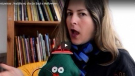 A imagem mostra a professora Carolina Hessel segurando um boneco de pano verde na frente de uma estante de livros. 