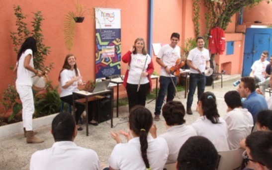 A foto retrata um grupo de alunos da Associação Nova Projeto sentados de frente para a equipe de educadores da Ritmos do Coração, que promovem uma interação por meio de instrumentos musicais como tambor, flauta transversal, violino e pratos.