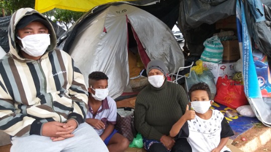 A imagem mostra um rapaz sentado em um banquinho, na rua, e, ao lado dele, três crianças e uma senhora de idade, sentadas no chão, na frente de uma barraca de camping, usando máscaras. 