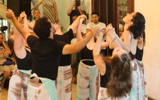 Um grupo de sete dançarinos fazem sua apresentação no salão da Casa das Rosas. Estão de mãos dadas e erguidas, olhando todos para cima. 